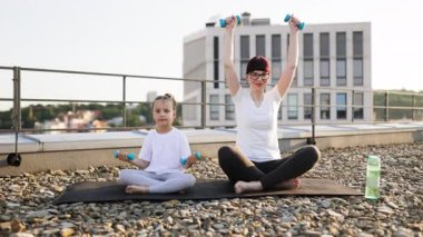 Anne ve kızı çatıda oturmuş dambıllarla egzersiz yapıyor, spor yapıyor, spor yapıyor ve kaynaşıyor. Parlak ve güneşli bir sabah, ikisi de sağlıklı yaşam tarzı aktiviteleriyle meşgul..