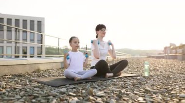 Anne ve kızı gün doğarken çatıda oturup halterlerle egzersiz yapıyorlar. Her iki model de spor kıyafetleri giyerek gülümsüyor. Aile spor kıyafetlerini, açık hava aktivitelerini destekliyor.