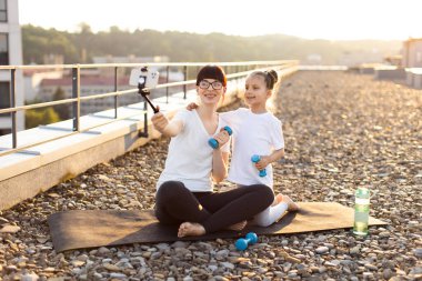 Anne ve küçük kızı damdaki dambıllarla yapılan spor salonunda selfie için poz veriyorlar. Parlak güneş ışığı neşeli atmosferi aydınlatıyor. Fitness bağı motivasyonunu ve sağlıklı bir yaşam tarzını gösteriyor..