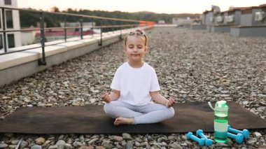 Nilüfer çiçeği pozisyonunda meditasyon yapan genç bir kız yanında ağırlık ve su şişesi de dahil olmak üzere günbatımında kentsel altyapıya sahip fitness objeleri var..