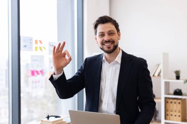 Takım elbiseli kendine güvenen yetişkin bir adam modern ofiste başarıyı simgeleyen iyi bir jest sergiliyor. Belgeler, klasörler ve dekorlarla görülebilen işyeri.