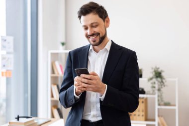 Resmi giyinmiş, gülümseyen ve modern ofiste akıllı telefon kullanan genç bir erkek. İletişim, iş ve profesyonelliği kendinden emin bir tavırla temsil eder..