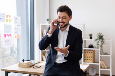 Takım elbiseli orta yaşlı bir işadamı parlak ofis yerinde telefonla konuşuyor, jestlerle gülümsüyor, modern çalışma ortamında.