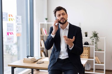 Takım elbiseli orta yaşlı bir işadamı parlak ofis yerinde telefonla konuşuyor, jestlerle gülümsüyor, modern çalışma ortamında.