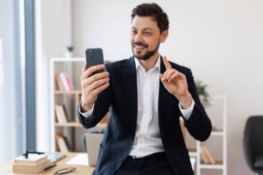 Beyaz takım elbiseli yetişkin bir adam kapalı alandaki akıllı telefondan görüntülü arama yapıyor. Ofis arka planda. İş ve iletişim kavramı.
