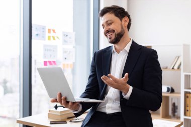 Resmi takım elbiseli profesyonel yetişkin bir erkek bilgisayar başında konuşuyor ve parlak modern ofis ortamında el kol hareketi yapıyor. Pozitif iletişim, dijital teknoloji ve iş kavramını yakalar.