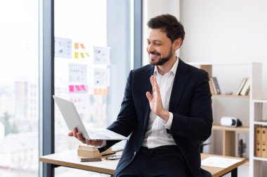Takım elbiseli bir iş adamı modern ofiste oturmuş internette el sallıyor, dizüstü bilgisayarla iletişim kuruyor. Pozitif ve profesyonel iş ortamı sergi teknolojisi