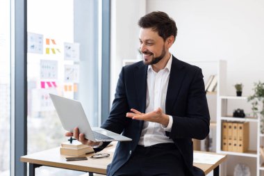 Takım elbiseli bir iş adamı modern ofiste oturmuş internette el sallıyor, dizüstü bilgisayarla iletişim kuruyor. Pozitif ve profesyonel iş ortamı sergi teknolojisi