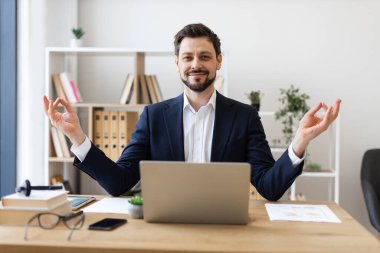 Profesyonel yetişkin erkek, Lotus 'ta ofis masasında oturmuş meditasyon yapıyor ve gülümsüyor, iş ortamında stres atmak ve bilinçlenmek için olumlu zihniyeti kucaklıyor.