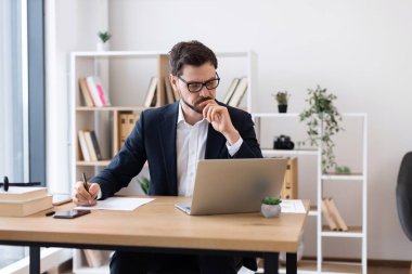 Takım elbiseli genç beyaz adam, organize ofis masasında çalışıyor, notlar yazıyor, dizüstü bilgisayar kullanıyor..