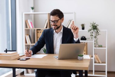 Genç beyaz erkek bilgisayar başında oturmuş profesyonel ofis ortamında notlar yazıyor. Raflı, dekorlu ve doğal ışıklandırmalı modern işyeri ortamı üretkenlik yaratıyor.