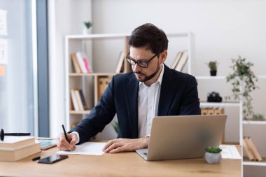 Genç beyaz erkek bilgisayar başında oturmuş profesyonel ofis ortamında notlar yazıyor. Raflı, dekorlu ve doğal ışıklandırmalı modern işyeri ortamı üretkenlik yaratıyor.