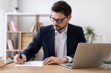 Resmi takım elbiseli genç, yetişkin bir erkek ofis düzeninde evrak işleriyle uğraşıyor. Masa ve dizüstü bilgisayarda iş ortamı, verimlilik, organizasyon vurgulandı.