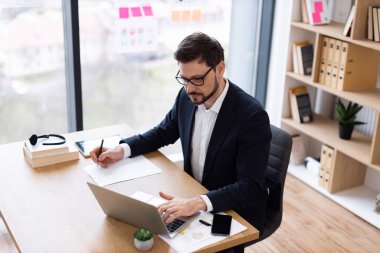 Resmi giyinmiş genç bir erkek, ofiste çalışıyor, dizüstü bilgisayar kullanıyor, kağıda not alıyor, iş yapıyor. Ofis ortamında doğal ışık, kitap rafları ve iş malzemeleri vardır..