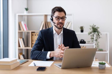 Yetişkin bir adam kulaklıkla takım elbise giyiyor. Modern ofiste video görüşmesi için dizüstü bilgisayar kullanıyor. Arkaplanda raflar ve bitkiler.