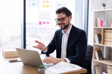 Modern ofis ortamında video görüşmesinde gülümseyen takım elbiseli profesyonel yetişkin erkek. Dizüstü bilgisayar masadayken kendinden emin bir hareket gösterir, uzaktan bağlantı kurar.