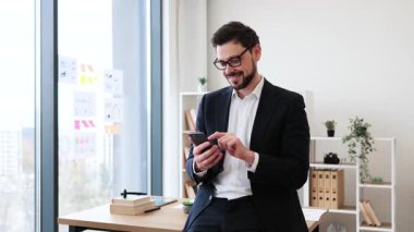 Resmi giyinmiş, gülümseyen ve modern ofiste akıllı telefon kullanan genç bir erkek. İletişim, iş ve profesyonelliği kendinden emin bir tavırla temsil eder..