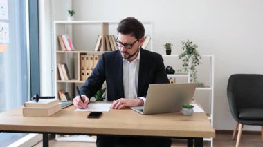 Ofis masasında dizüstü bilgisayarla çalışan resmi giyinmiş genç bir erkek not yazıyor. Modern çalışma alanı, verimli ve profesyonel atmosferi gösteren belgeler, kitaplar ve saksı bitkilerini içerir.
