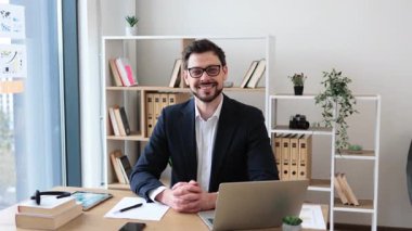 Resmi takım elbiseli gülümseyen beyaz bir erkek dizüstü bilgisayar ve kağıtlarla çalışıyor. Profesyonel, üretken ve odaklanmış iş dünyası modern ofis atmosferini gözler önüne seriyor..