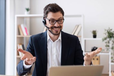 Ofisteki video görüşmesi sırasında kulaklık ve dizüstü bilgisayar kullanan resmi takım elbiseli gülümseyen genç adam. Arka planda kitaplıkları olan profesyonel çalışma ortamı uzak çalışma için neşeli yaklaşımlar sergiliyor.