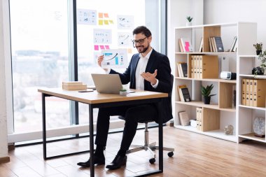 Takım elbiseli genç beyaz erkek, parlak modern iş yerinde grafik ve çizelge kullanarak masa başında oturuyor. Ofis arkaplanı kitaplı ve dosyalı rafları içerir.