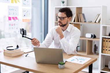 Profesyonel iş adamı, 30 'lu yaşlarda, gözlüklü, ofis masasında oturmuş video görüşmesi yapan ya da akıllı telefonlu video bloğu olan. Arkaplanda raflar olan parlak modern çalışma alanı