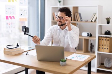 Profesyonel iş adamı, 30 'lu yaşlarda, gözlüklü, ofis masasında oturmuş video görüşmesi yapan ya da akıllı telefonlu video bloğu olan. Arkaplanda raflar olan parlak modern çalışma alanı