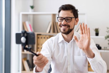 Orta yaşlı bir adam akıllı telefonuyla video kaydediyor. Mutluluk ve nişanı ifade ediyor. Beyaz gömlekli, modern ofis ortamında yakalanmış..