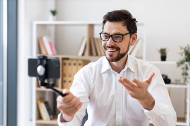 Orta yaşlı bir adam akıllı telefonuyla video kaydediyor. Mutluluk ve nişanı ifade ediyor. Beyaz gömlekli, modern ofis ortamında yakalanmış..
