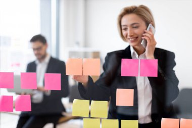 Cam tahtası olan genç bir iş kadını proje planlaması için yapışkan notlar kullanıyor, iş arkadaşı çalışıyor. Ofis, odak, işbirliği, organize çalışma stratejisini gösteren modern çalışma ortamı