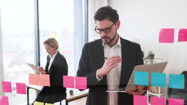 İş adamı cam tahtaya yapışkan notlarla fikirler üzerinde çalışıyor. Arka planda da iş kadını. İşbirlikçi takım, modern ofis ortamları, beyaz erkek ve kadın profesyoneller..