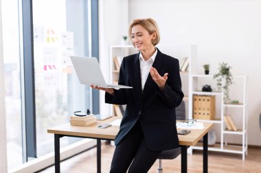 Kendine güvenen orta yaşlı beyaz kadın iş kıyafeti giymiş ofis ortamında video görüşmesi yapıyor. Profesyonel çalışma alanı temel gereksinimlere, büyük pencerelere ve organize raflara sahip masaları içerir..