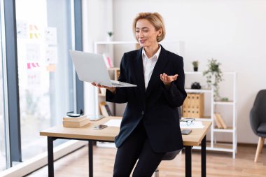 Kendine güvenen orta yaşlı beyaz kadın iş kıyafeti giymiş ofis ortamında video görüşmesi yapıyor. Profesyonel çalışma alanı temel gereksinimlere, büyük pencerelere ve organize raflara sahip masaları içerir..