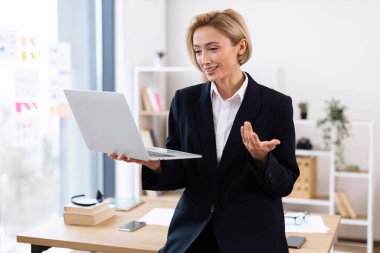 Video konferansı sırasında dizüstü bilgisayar kullanarak resmi giyinmiş kendinden emin genç bir iş kadını. Kitap rafları ve kağıtları ile ofis çalışma alanı arka planı profesyonel iletişim ayarlarını öneriyor