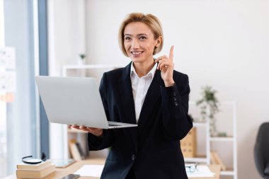 Ofiste kendine güvenen orta yaşlı bir iş kadını dizüstü bilgisayar tutuyor, gülümsüyor, fikir veriyor, resmi kıyafetler giyiyor. Masa ve arkaplan dekorlu profesyonel çalışma alanı dinamik ortam sağlar.