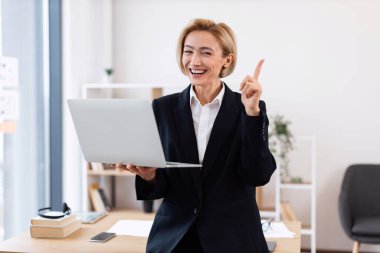 Ofiste kendine güvenen orta yaşlı bir iş kadını dizüstü bilgisayar tutuyor, gülümsüyor, fikir veriyor, resmi kıyafetler giyiyor. Masa ve arkaplan dekorlu profesyonel çalışma alanı dinamik ortam sağlar.