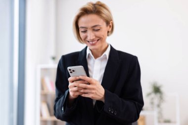 Kendine güvenen yetişkin iş kadını resmi siyah takım elbise giyiyor. Parlak ofis alanında çalışırken akıllı telefon kullanıyor.