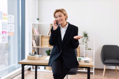 30 'lu yaşlarda kendine güvenen, resmi giyinmiş, şık ofis ortamında telefonla konuşan, arka planda masası ve rafları olan, iş başarısını ve iletişimini aktaran bir kadın.