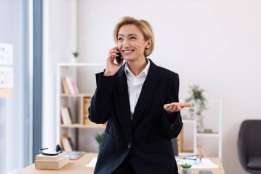 30 'lu yaşlarda kendine güvenen, resmi giyinmiş, şık ofis ortamında telefonla konuşan, arka planda masası ve rafları olan, iş başarısını ve iletişimini aktaran bir kadın.