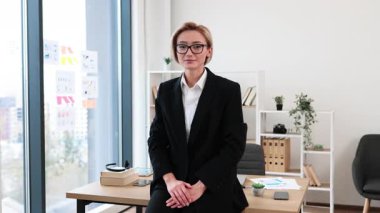 Profesyonel, takım elbiseli genç bir kadın masasında kendine güvenerek duruyor. Parlak ofis ortamında, profesyonelliği ve başarıyı temsil ediyor..