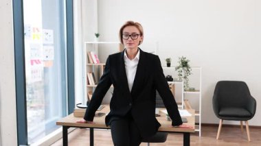 Modern ofiste kendine güveni olan profesyonel bir kadın resmi takım elbise giyiyor. Masası, bitkileri ve arka planda rafları olan iş yeri. Başarı, profesyonellik ve motivasyonu temsil eder