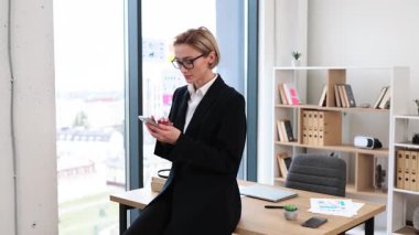 Modern ofisteki profesyonel iş kadını masaüstü telefonuna yaslanıyor, resmi kıyafetler giyiyor. Rafları ve cam pencereleri olan parlak bir iş yeri. Üretkenliği, iletişimi, çoklu görevi temsil eder