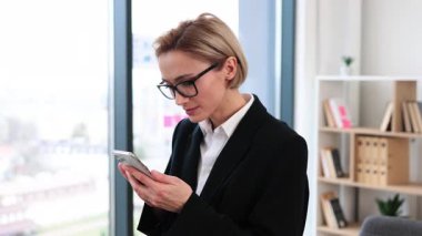 Kendine güveni tam orta yaşlı bir kadın resmi giyinmiş parlak bir ofiste elinde akıllı bir telefonla gülümsüyor.