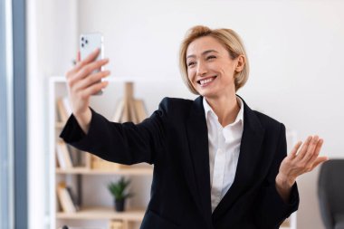 Resmi siyah takım elbiseli gülümseyen bir iş kadını. Parlak ofis ortamında uzaktan video görüşmesi yapıyor. İletişim, teknoloji ve profesyonel etkileşim kavramlarının temsili.
