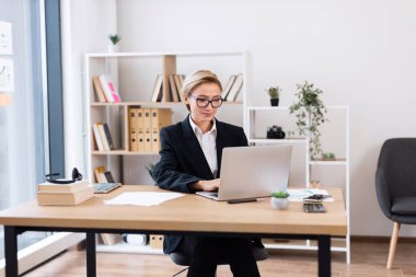 Siyah takım elbiseli iş kadını parlak ofis ortamında dizüstü bilgisayar kullanarak masada oturuyor. Yetişkin kadın olarak tasvir edilen ofis çalışanı proje uygulamasına odaklandı.