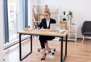 Otuzlarında profesyonel bir kadın modern ofiste masa başında oturmuş belgeleri analiz ediyor ve dizüstü bilgisayarla çalışıyor. Çalışma alanı, üretkenliği ve odaklanmayı artıran doğal ışık, organize raflar içerir..