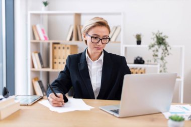 Orta yaşlı, gözlüklü, ofiste dizüstü bilgisayarla kağıt üzerinde çalışan sarışın bir kadın..