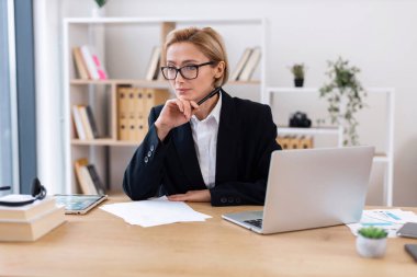 Gözlüklü ve sarı saçlı bir iş kadını dizüstü bilgisayarla masada oturuyor. Ofisteki orta yaşlı profesyoneller işe odaklandılar. Liderlik kavramı verimlilik kariyeri ve işyeri.