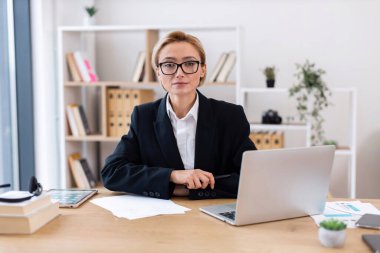 Modern ofis ortamında çalışan kendine güvenen, resmi giyinmiş yetişkin bir kadın. İşletmeyi, profesyonelliği ve üretkenliği vurgulayan dizüstü bilgisayarlı, kağıtlı, kitaplı profesyonel odaklanma. Parlak çalışma alanı.