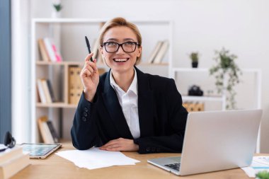 Ofis masasında dizüstü bilgisayarla oturan kendine güvenen yetişkin bir kadın. Kadın resmi kıyafetler giyiyor, modern, düzenli çalışma alanında çalışırken kendine güvenerek gülümsüyor..